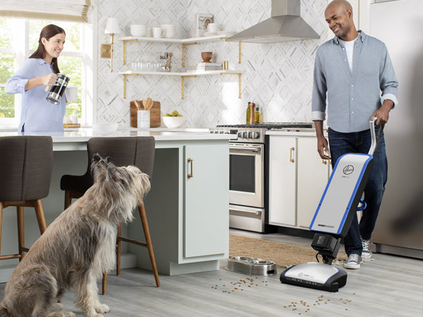 Man vacuuming dog food off of hardwood floor with woman and dog in background 